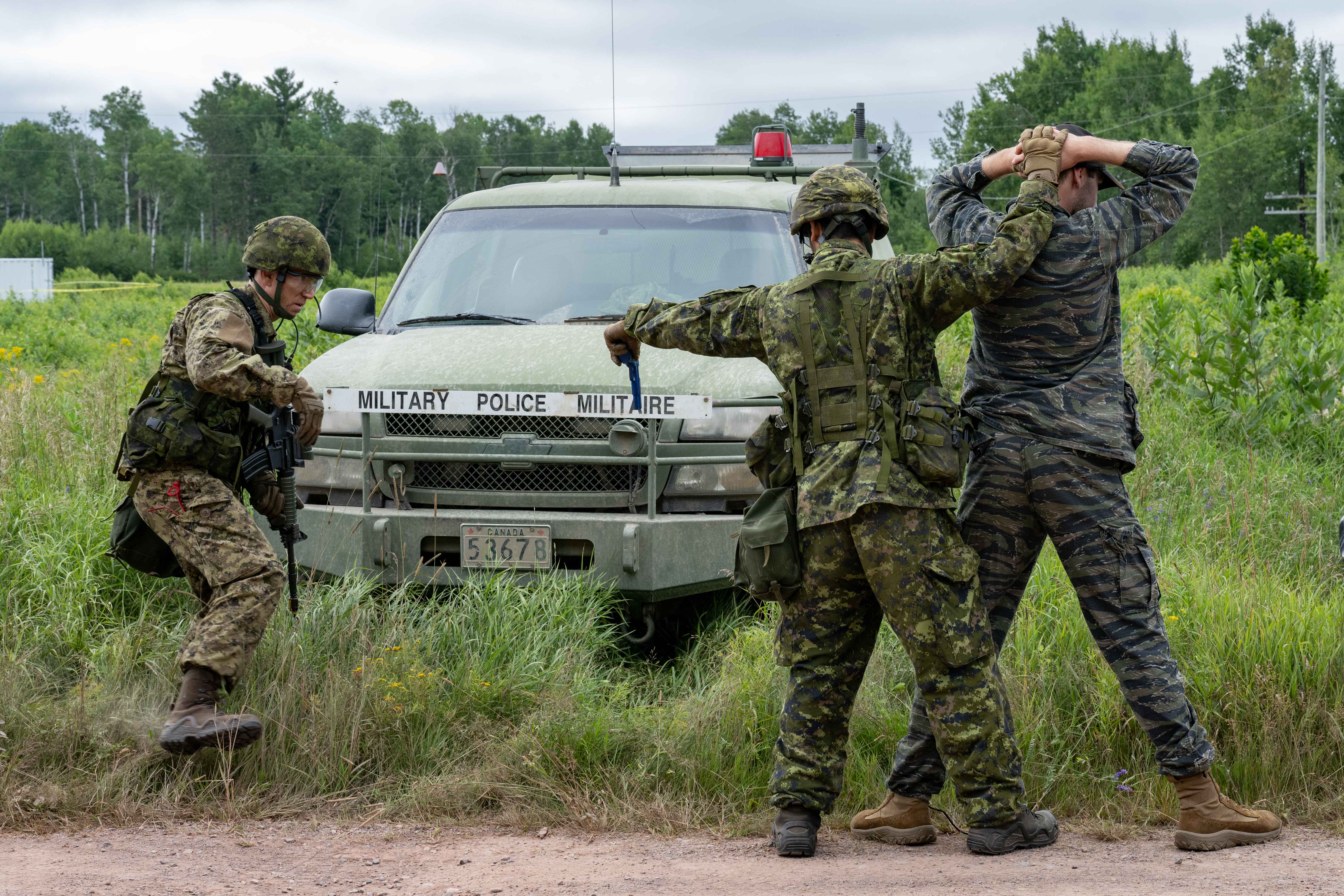 Two Miliary Police members dressed in camouflage detain a person in a dark uniform during a
training exercise. The detainee stands with hands placed behind their head, while a green
Military Police truck is visible in the background, parked on a grassy field.